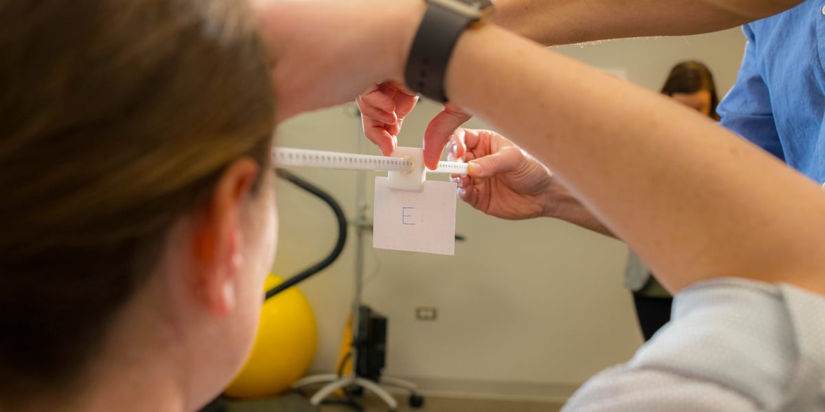 A patient (back side of head only) undergoes a visual test. A provider (arm only) slides a card with a capital E farther away from the patient's eyes.
